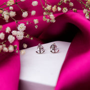 A pair of silver stud earrings designed to resemble an anchor, displayed on a pink fabric backdrop with decorative flowers around.